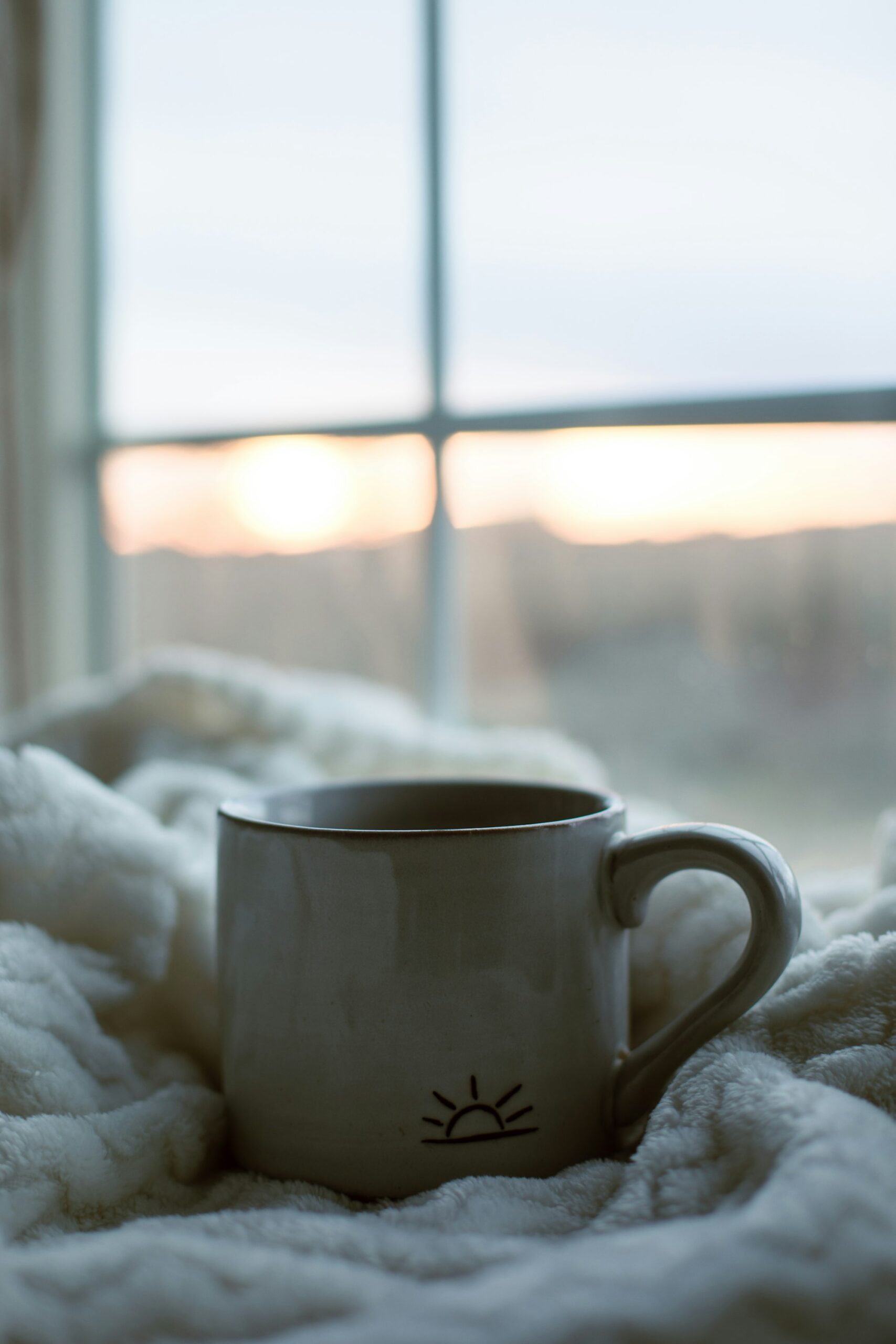 A ceramic cup on a white blanket in front of a window at sunrise