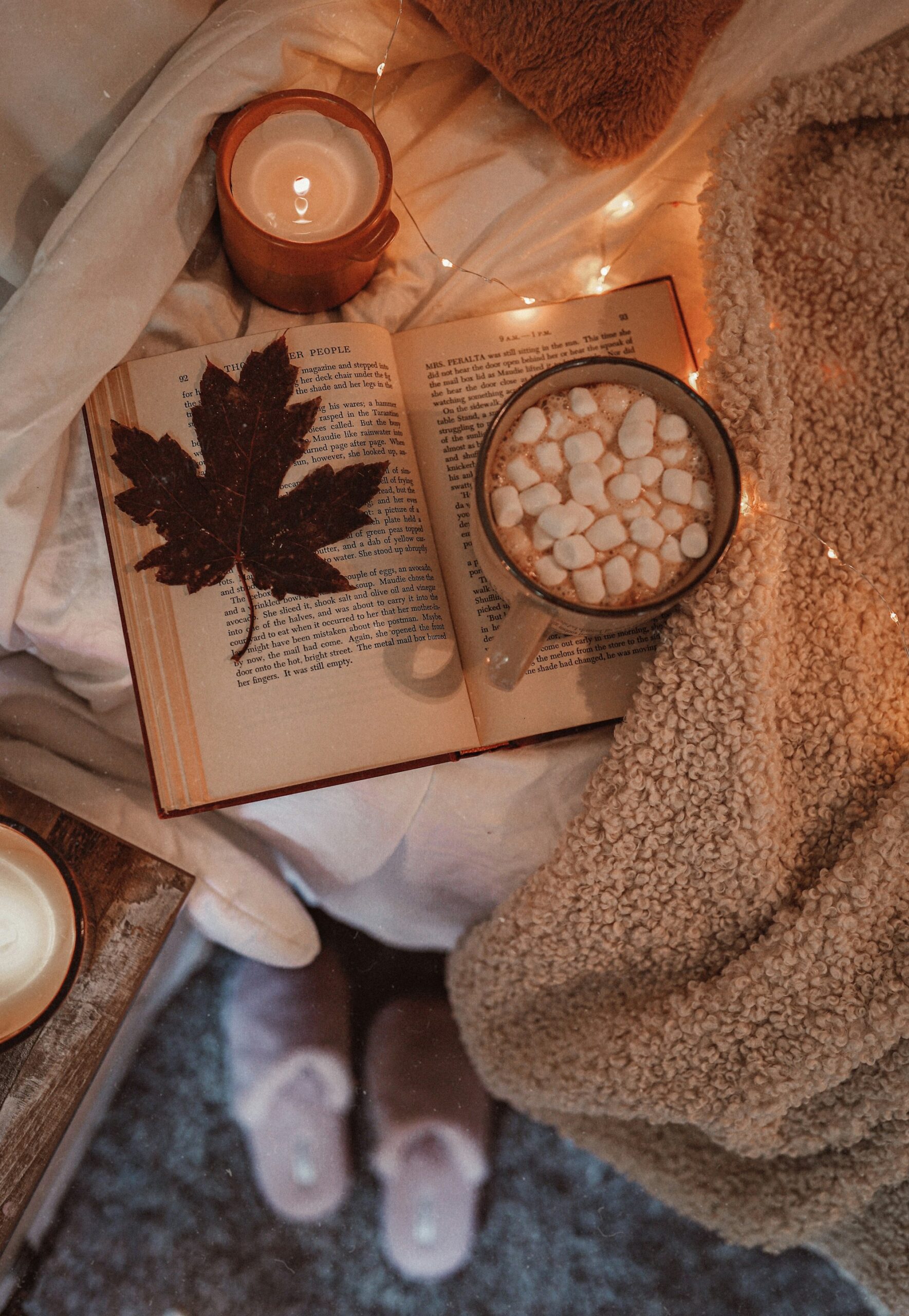 An open book with a maple leaf and a bowl of marshmallows on a knitted throw